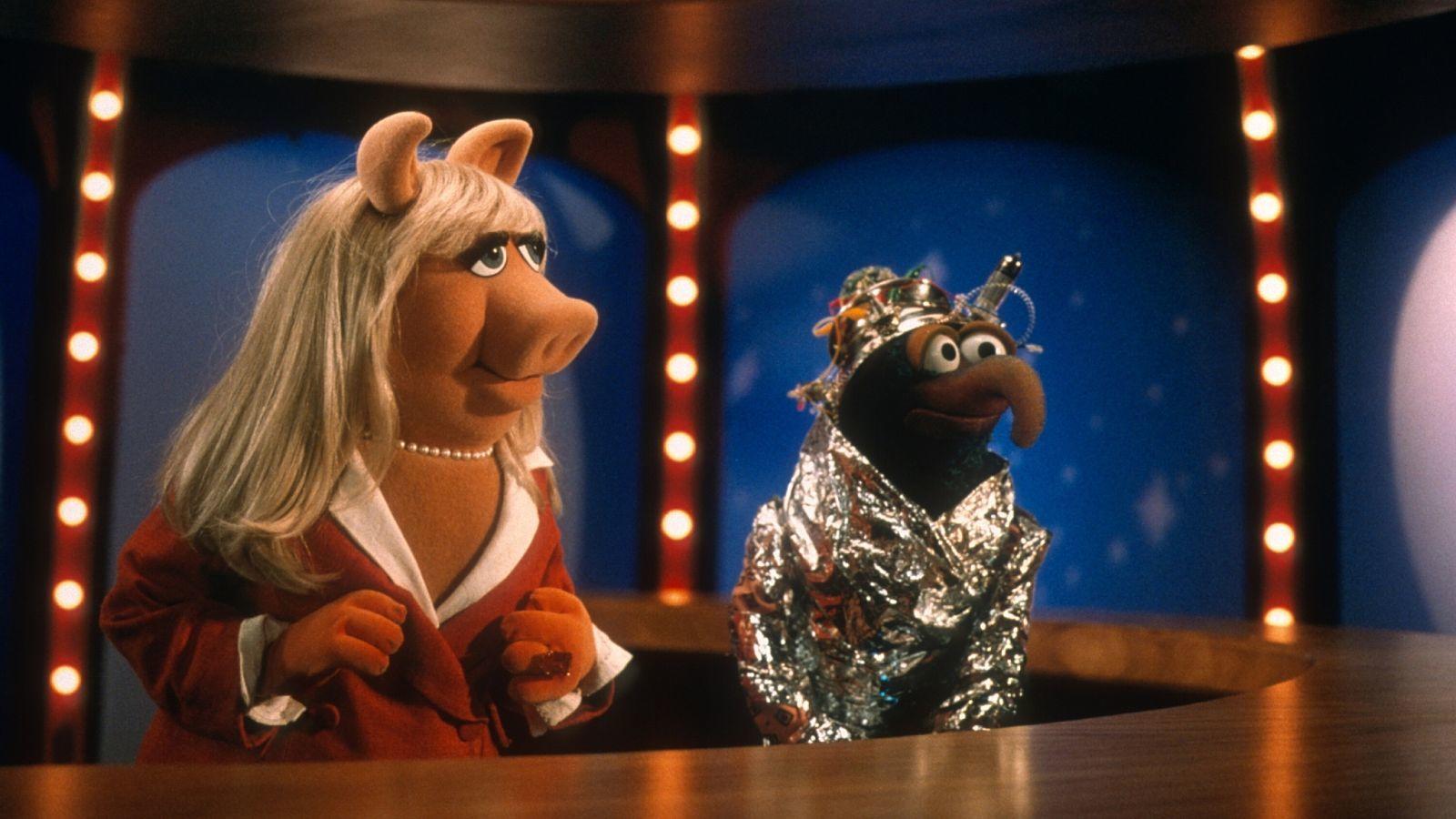 screenshot from 'Muppets from Space' showing Miss Piggy as a news anchor next to Gonzo who is wrapped in tin foil and wearing a strange scientific device on his head