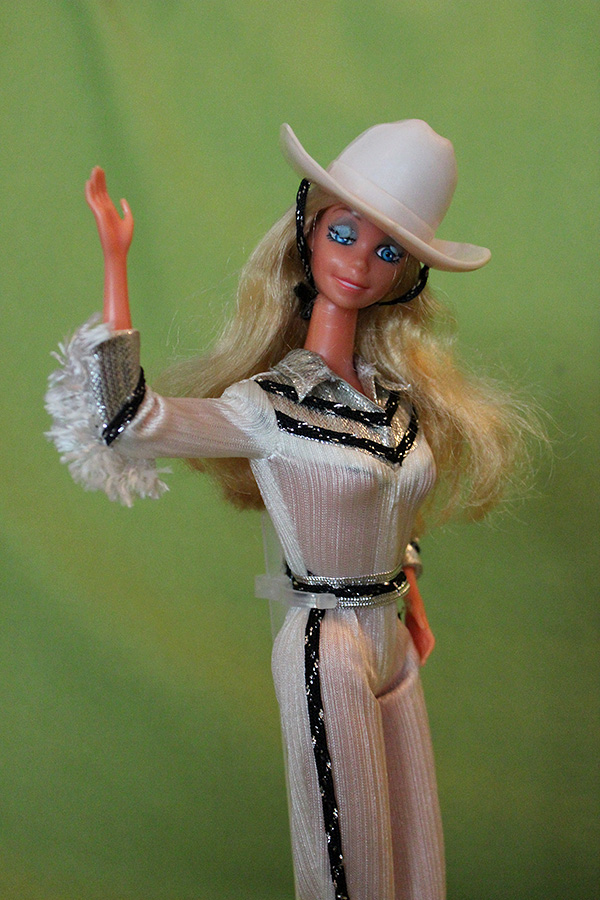 a photo of Western Barbie in her white cowgirl outfit with one hand up as if she is waving and a half-wink expression on her face